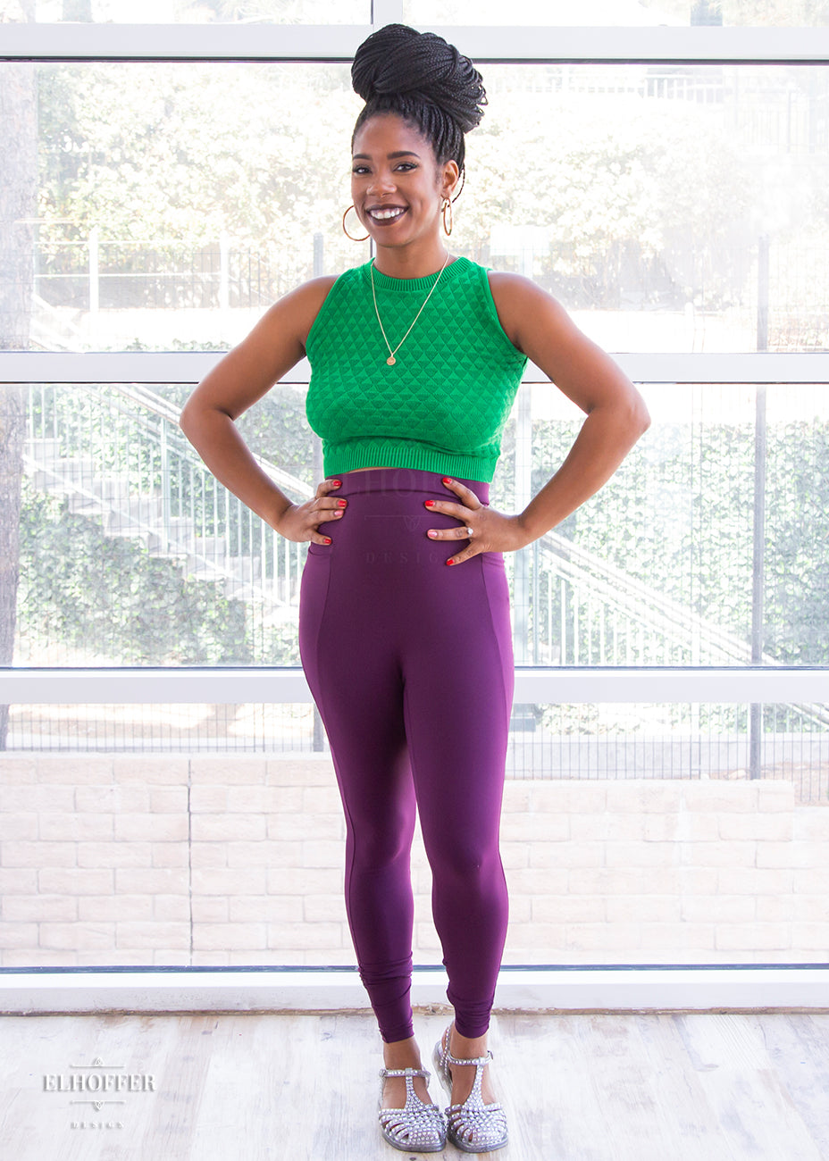 Krystina, a size small medium dark skinned model with long braids, is wearing a pair of high waisted leggings with a seamless front and side seam pockets in plum.