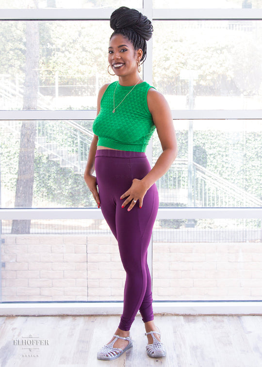 Krystina, a size small medium dark skinned model with long braids, is wearing a pair of high waisted leggings with a seamless front and side seam pockets in plum.