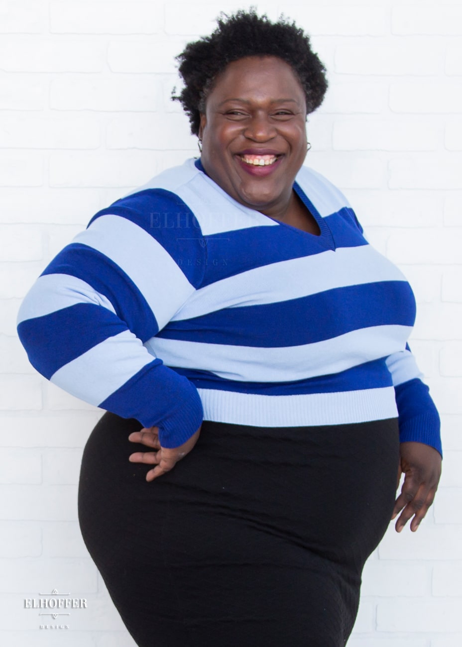 Adalgiza, a medium dark skinned 4xl model with short black super curly hair, is smiling while wearing a cropped v-neck sweater with fitted full length sleeves in horizontal thick stripes of bright blue and light blue.