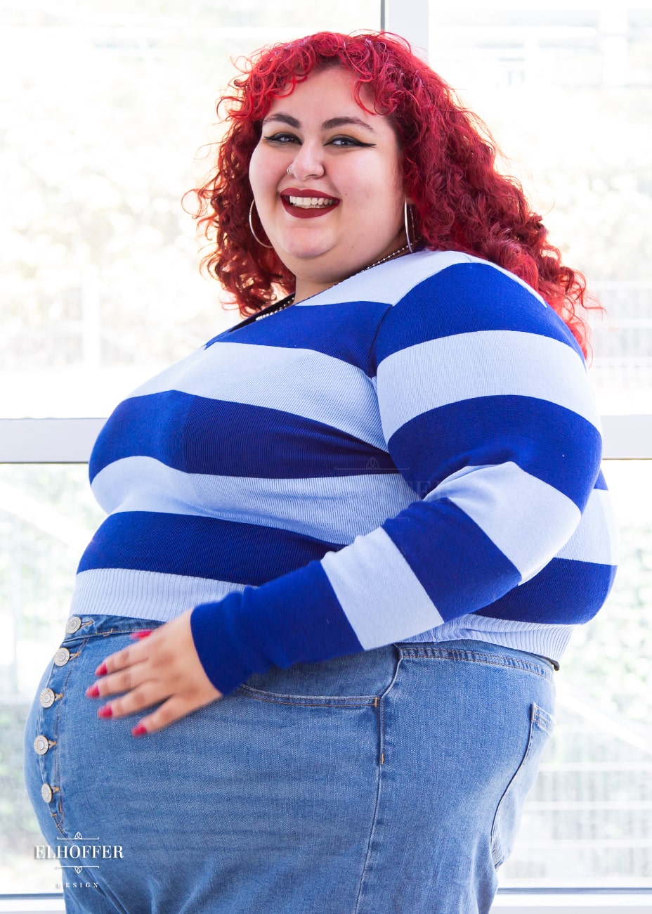 Victoria, an olive skinned size 4XL model with bright red curly hair, is wearing a cropped v-neck sweater with fitted full length sleeves in horizontal thick stripes of bright blue and light blue.