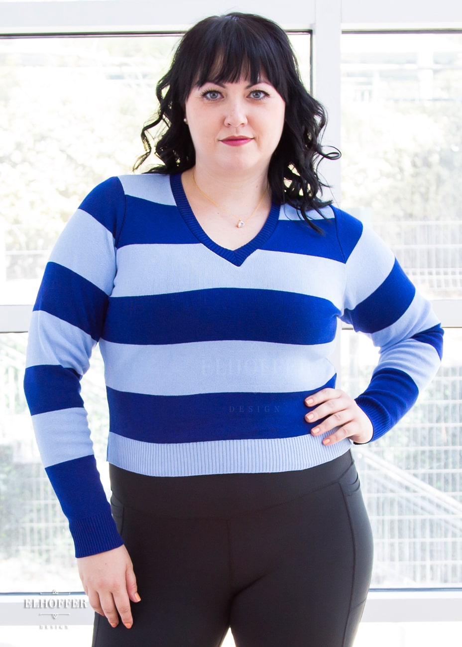Bernadette, a fair skinned size large model with short black hair and bangs,  is wearing a cropped v-neck sweater with fitted full length sleeves in horizontal thick stripes of bright blue and light blue.