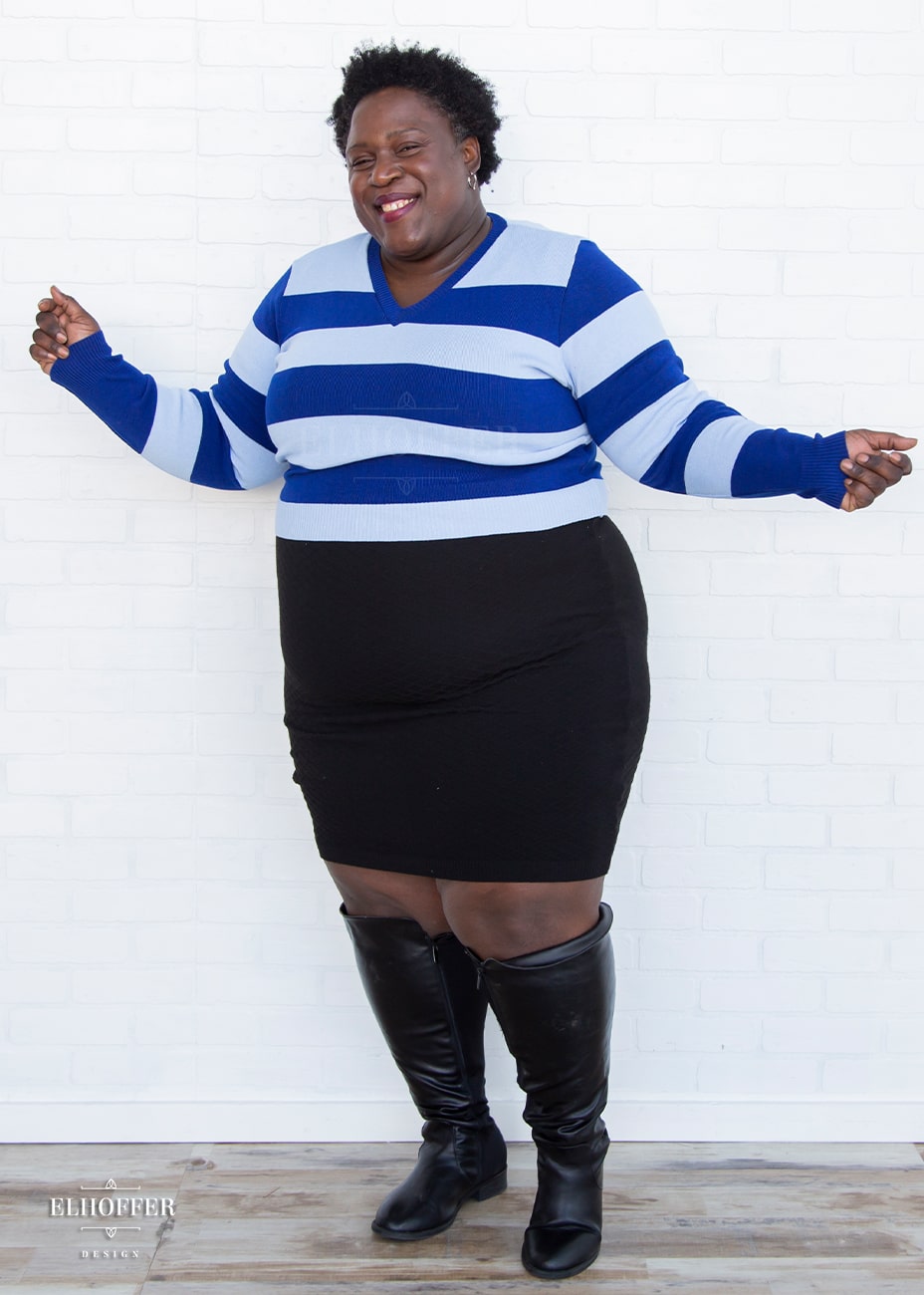 Adalgiza, a medium dark skinned 4xl model with short black super curly hair, is smiling while wearing a cropped v-neck sweater with fitted full length sleeves in horizontal thick stripes of bright blue and light blue.