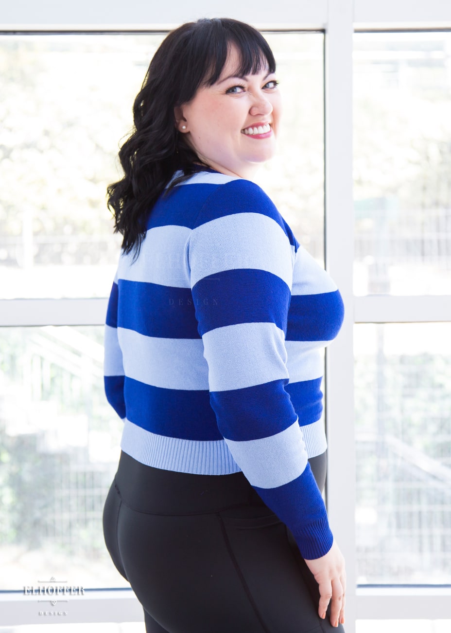 Bernadette, a fair skinned size large model with short black hair and bangs, is wearing a cropped v-neck sweater with fitted full length sleeves in horizontal thick stripes of bright blue and light blue.