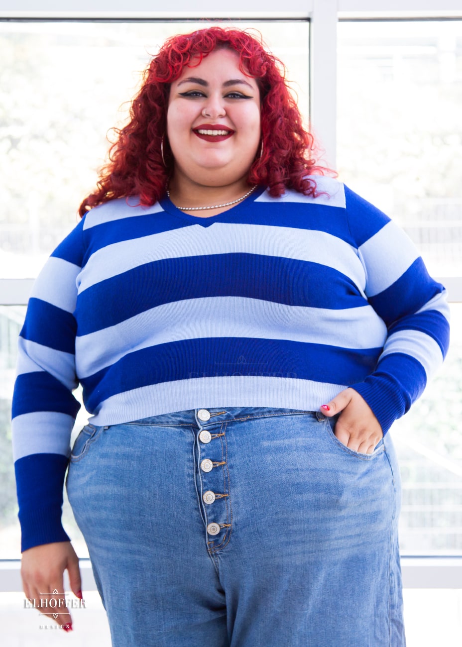 Victoria, an olive skinned size 4XL model with bright red curly hair, is wearing a cropped v-neck sweater with fitted full length sleeves in horizontal thick stripes of bright blue and light blue.