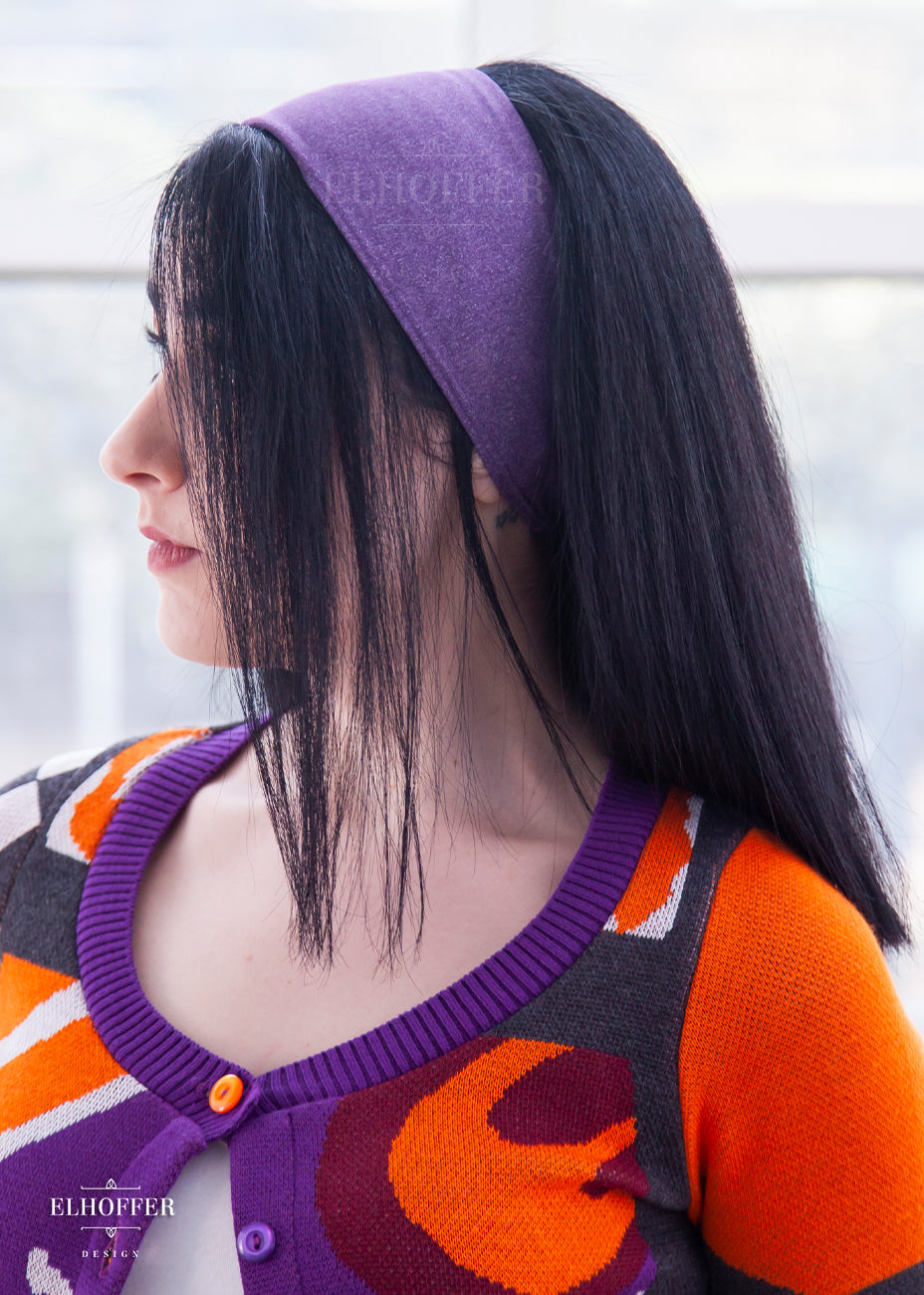 A side view of a heathered purple headband with hemmed edges. The headband is around 3" at the top and gradually gets smaller as it goes around the head.