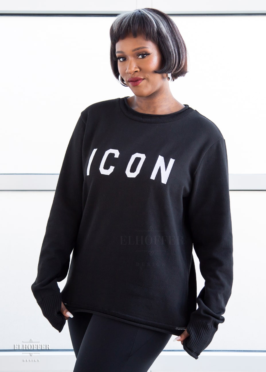 Lynsi, a medium dark skinned M model with short black and white hair, is wearing an XL sample of black unisex sweater with white bold embroidered letters that spell ICON and has long sleeves with thumbholes. The sweater also has a rolled hem on both the neckline and around the cuffs. The XL sample makes the sweater look like a relaxed fit, she would normally wear a M, or a S if she wanted it more fitted.