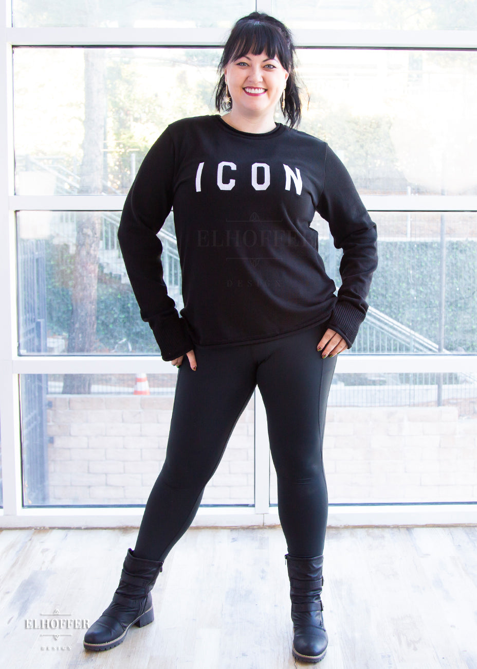 Bernadette, a fair skinned L model with long dark brown hair pulled up in a pony tail and bangs, is smiling while wearing an XL sample of black unisex sweater with white bold embroidered letters that spell ICON and has long sleeves with thumbholes. The sweater also has a rolled hem on both the neckline and around the cuffs. The XL sample makes the sweater look like a relaxed fit, she would get her normal L size if she wanted it more fitted.