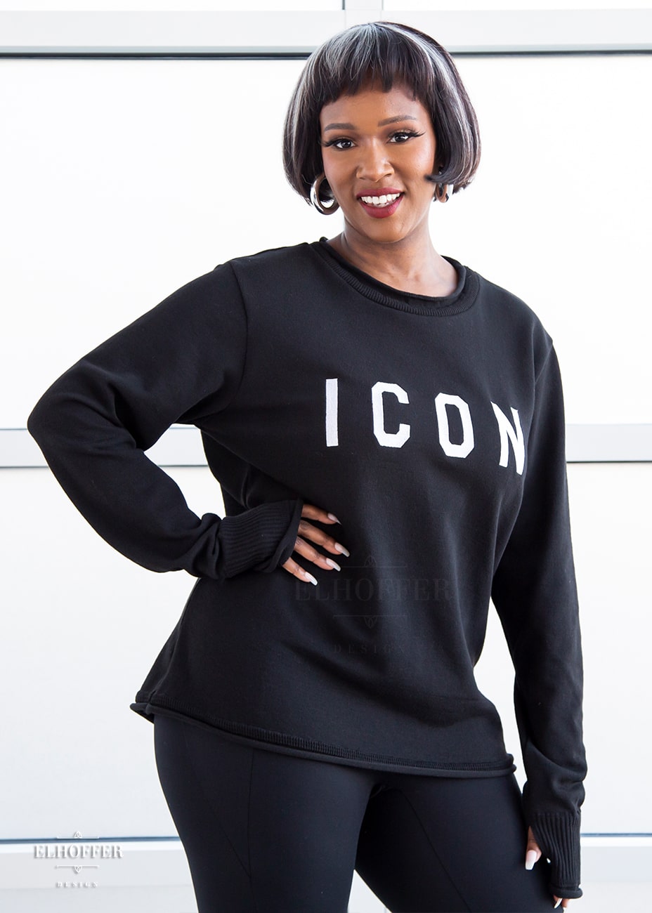 Lynsi, a medium dark skinned M model with short black and white hair, is smiling while wearing an XL sample of black unisex sweater with white bold embroidered letters that spell ICON and has long sleeves with thumbholes. The sweater also has a rolled hem on both the neckline and around the cuffs. The XL sample makes the sweater look like a relaxed fit, she would normally wear a M, or a S if she wanted it more fitted.
