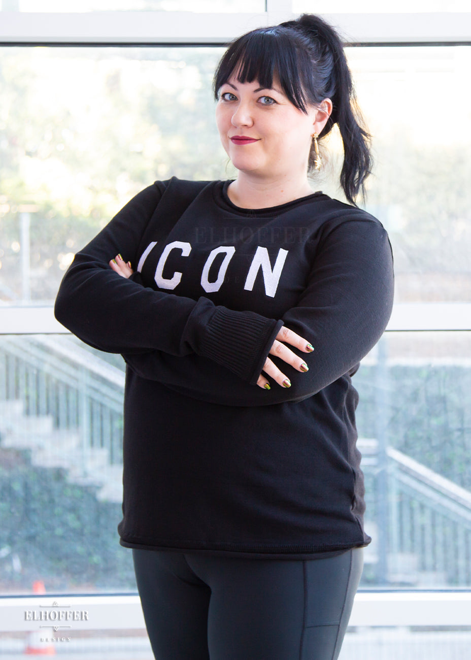 Bernadette, a fair skinned L model with long dark brown hair pulled up in a pony tail and bangs, is wearing an XL sample of black unisex sweater with white bold embroidered letters that spell ICON and has long sleeves with thumbholes. The sweater also has a rolled hem on both the neckline and around the cuffs. The XL sample makes the sweater look like a relaxed fit, she would get her normal L size if she wanted it more fitted.