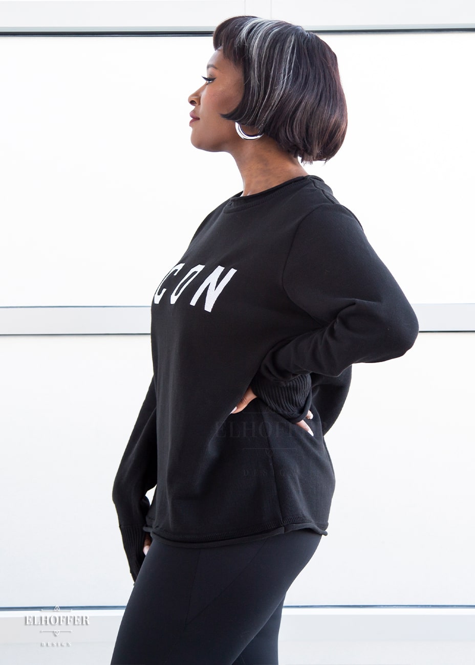 A side view of Lynsi, a medium dark skinned M model with short black and white hair, wearing an XL sample of black unisex sweater with white bold embroidered letters that spell ICON and has long sleeves with thumbholes. The sweater also has a rolled hem on both the neckline and around the cuffs. The XL sample makes the sweater look like a relaxed fit, she would normally wear a M, or a S if she wanted it more fitted.