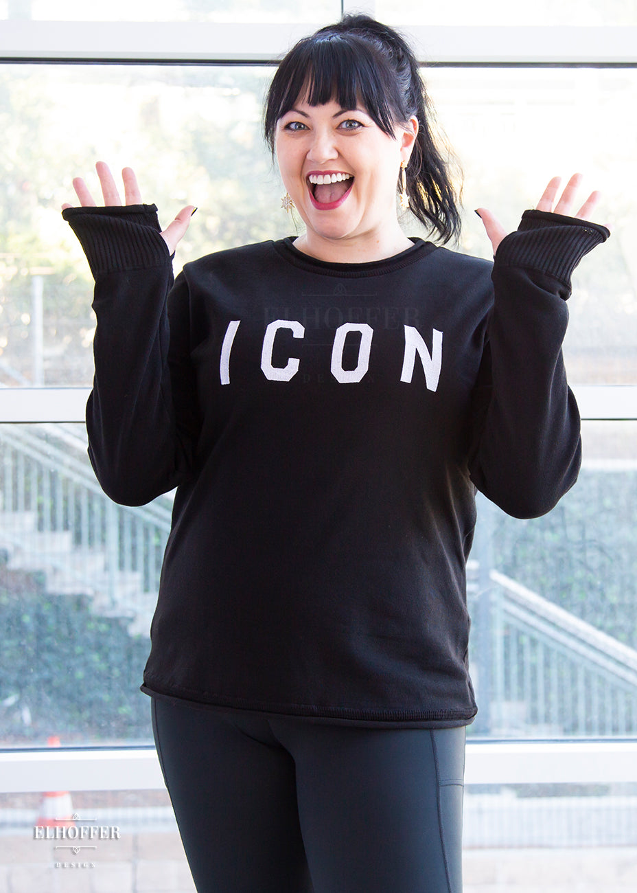 Bernadette, a fair skinned L model with long dark brown hair pulled up in a pony tail and bangs, is smiling with her hands up to show off the thumbholes in the cuffs of the XL sample of a black unisex sweater that has white bold embroidered letters that spell ICON and has long sleeves. The sweater also has a rolled hem on both the neckline and around the cuffs. The XL sample makes the sweater look like a relaxed fit, she would get her normal L size if she wanted it more fitted.