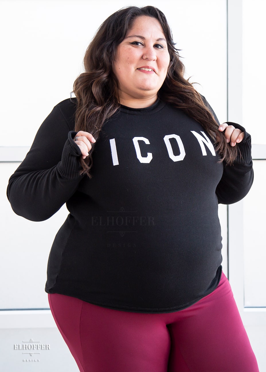 Alysia, a sun kissed skin 2xl model with long dark brown wavy hair, is smiling while wearing an XL sample of black unisex sweater with white bold embroidered letters that spell ICON and has long sleeves with thumbholes. The sweater also has a rolled hem on both the neckline and around the cuffs. The XL gives her a more fitted look, if she wanted a more relaxed look, she would wear her normal size of 2xl.