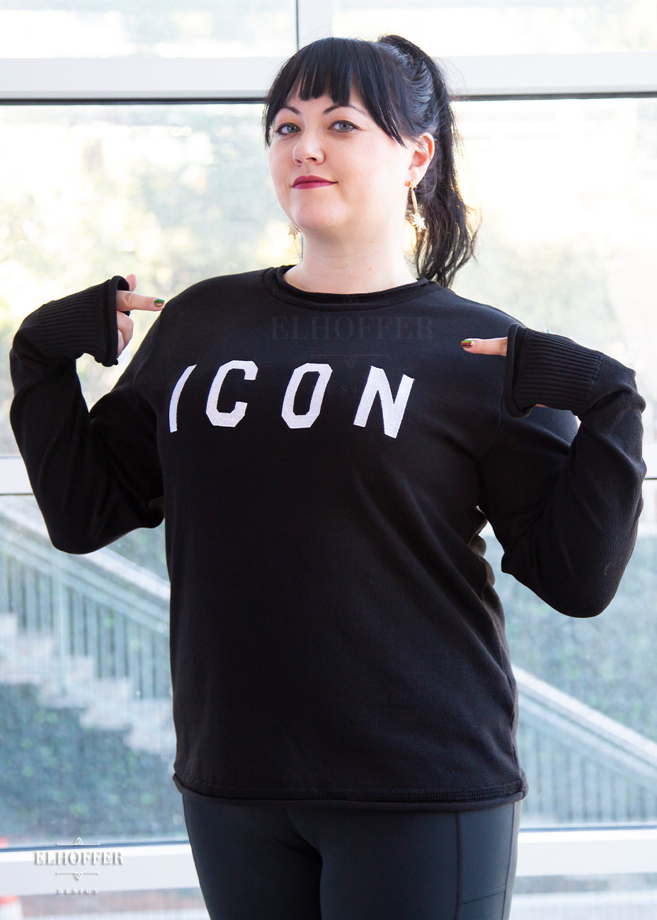 Bernadette, a fair skinned L model with long dark brown hair pulled up in a pony tail and bangs, is wearing an XL sample of black unisex sweater with white bold embroidered letters that spell ICON and has long sleeves with thumbholes. The sweater also has a rolled hem on both the neckline and around the cuffs. The XL sample makes the sweater look like a relaxed fit, she would get her normal L size if she wanted it more fitted.