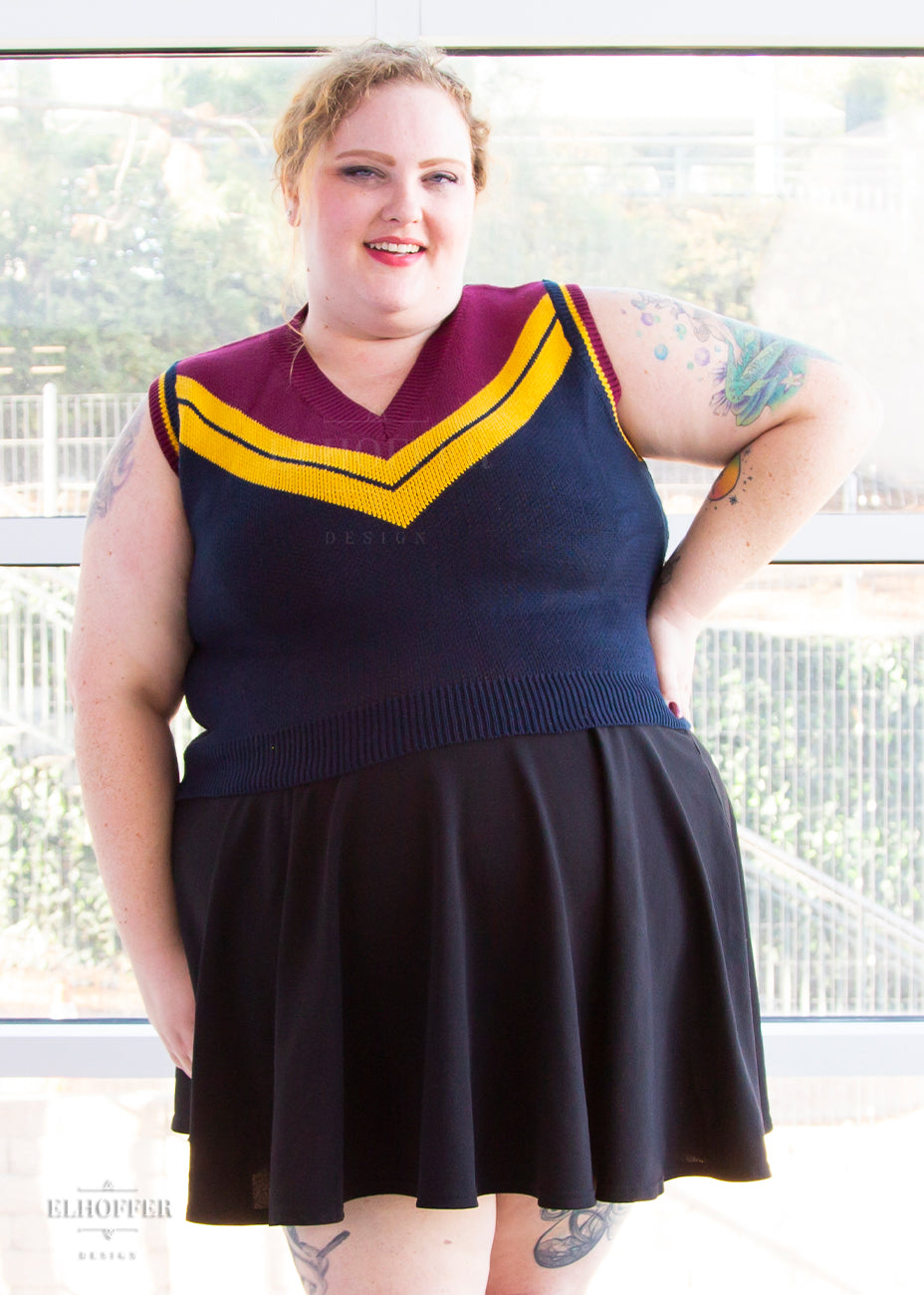 Bee, a size 3XL fair skinned model with ginger hair, is wearing a pullover vest with a boxy fit and v-neck. It is navy blue with a deep red neckline and mustard yellow v detail.