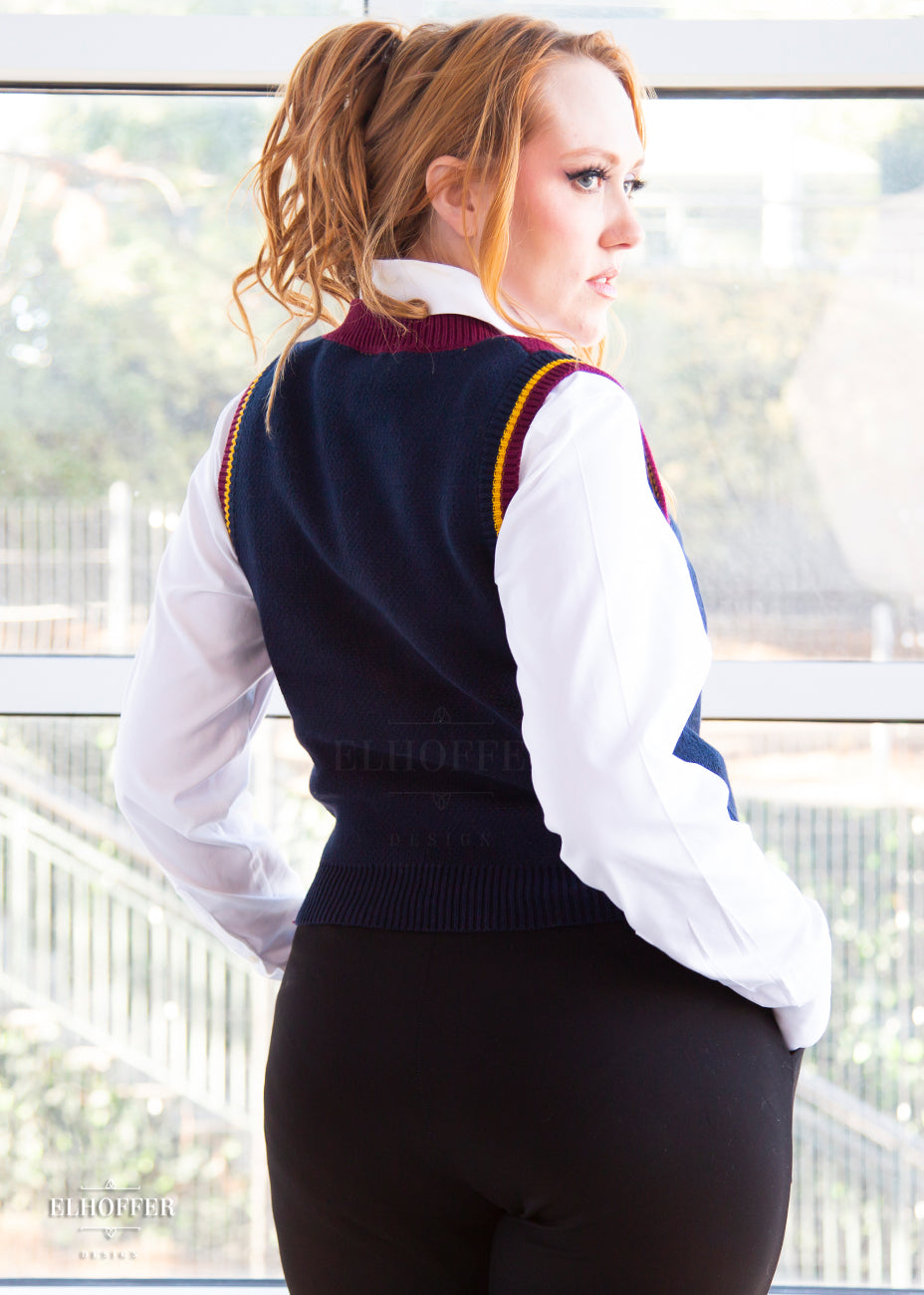 Kelsey, a size small model with long ginger hair, is wearing a pullover vest with a boxy fit and v-neck. It is navy blue with a deep red neckline and mustard yellow v detail. The back, shown here, is navy except for the deep red neckline and mustard and deep red arm cuffs.