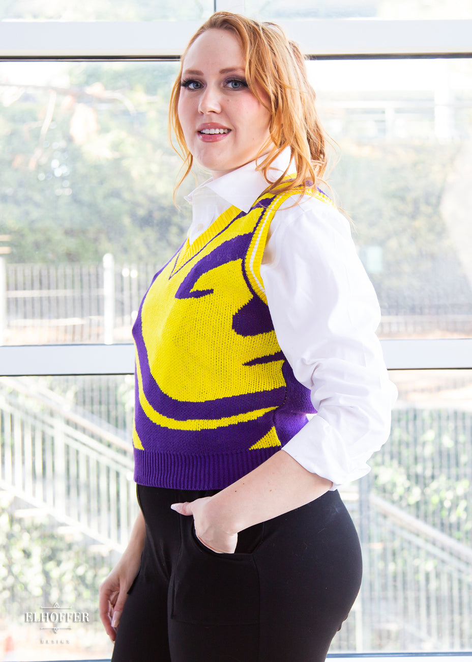 Kelsey, a size small fair skinned model with long ginger hair, is wearing a pullover vest with a boxy fit and v-neck. It is bright purple with a yellow swirl detail across the body.