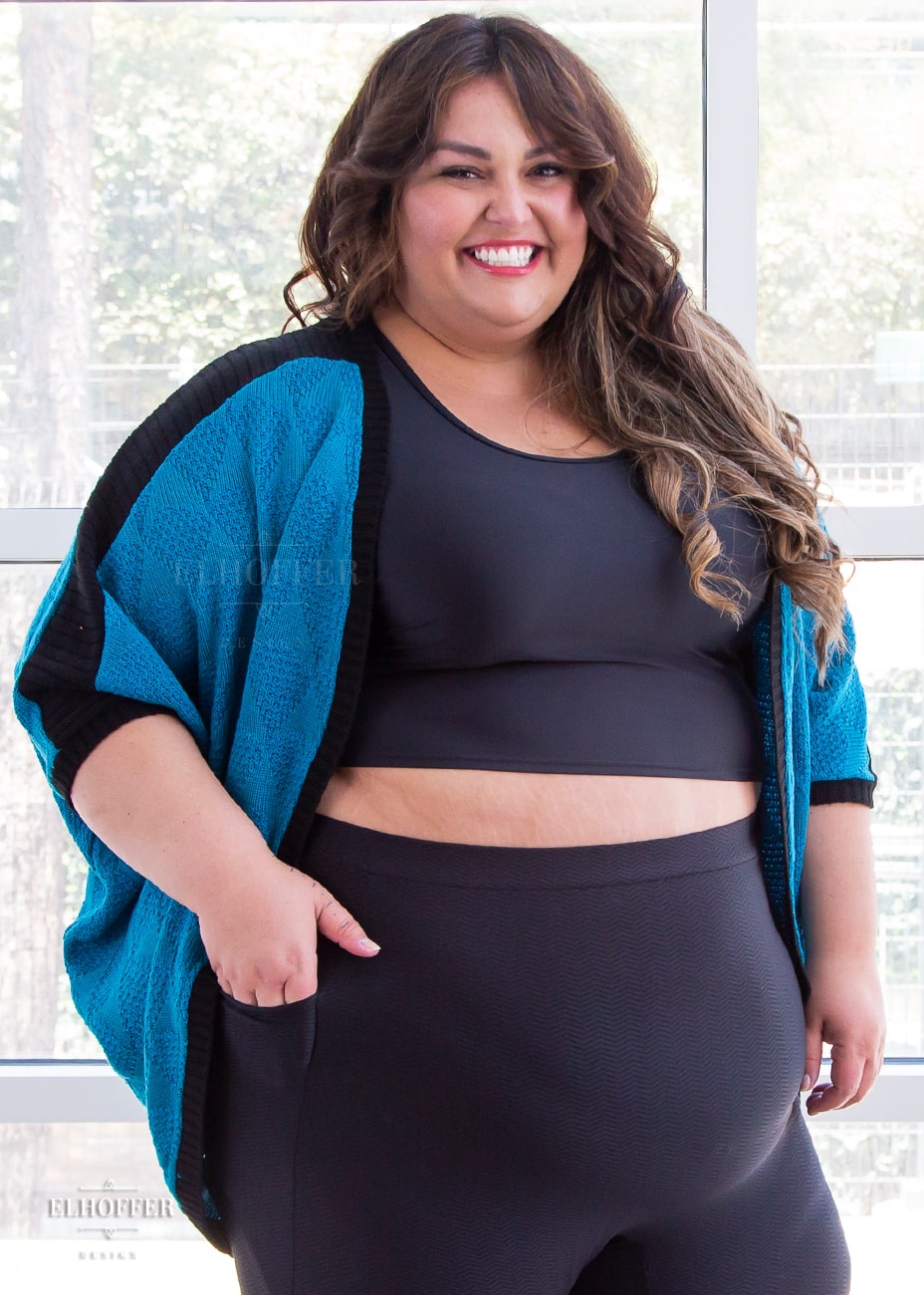 Kristen, a size 3XL olive skinned model with long highlighted brown hair, is wearing a teal open front dolman shrug featuring black ribbed shoulder detailing.