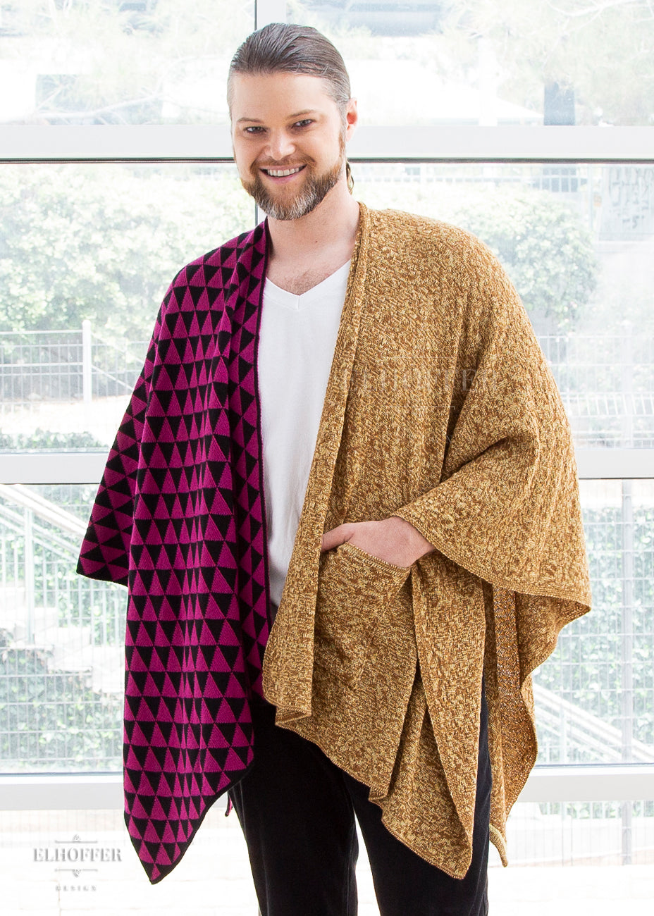 Bryan, a fair skinned and blonde haired size L model with facial hair, wears the one size poncho. One side of the poncho is a black and pink triangle pattern and the other side is a mustard blend of yarn with a pocket. He has paired it with black jeans and a white t shirt.