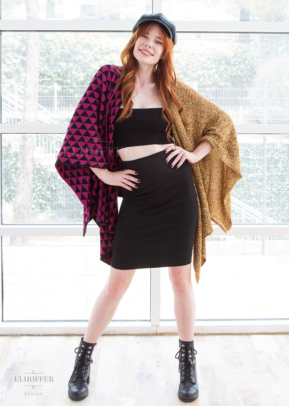 Chloe, a fair skinned ginger haired size XS model, wears the poncho with her hands on her hips. She pairs it with a black crop top, black bodycon skirt, black boots, and a black hat.