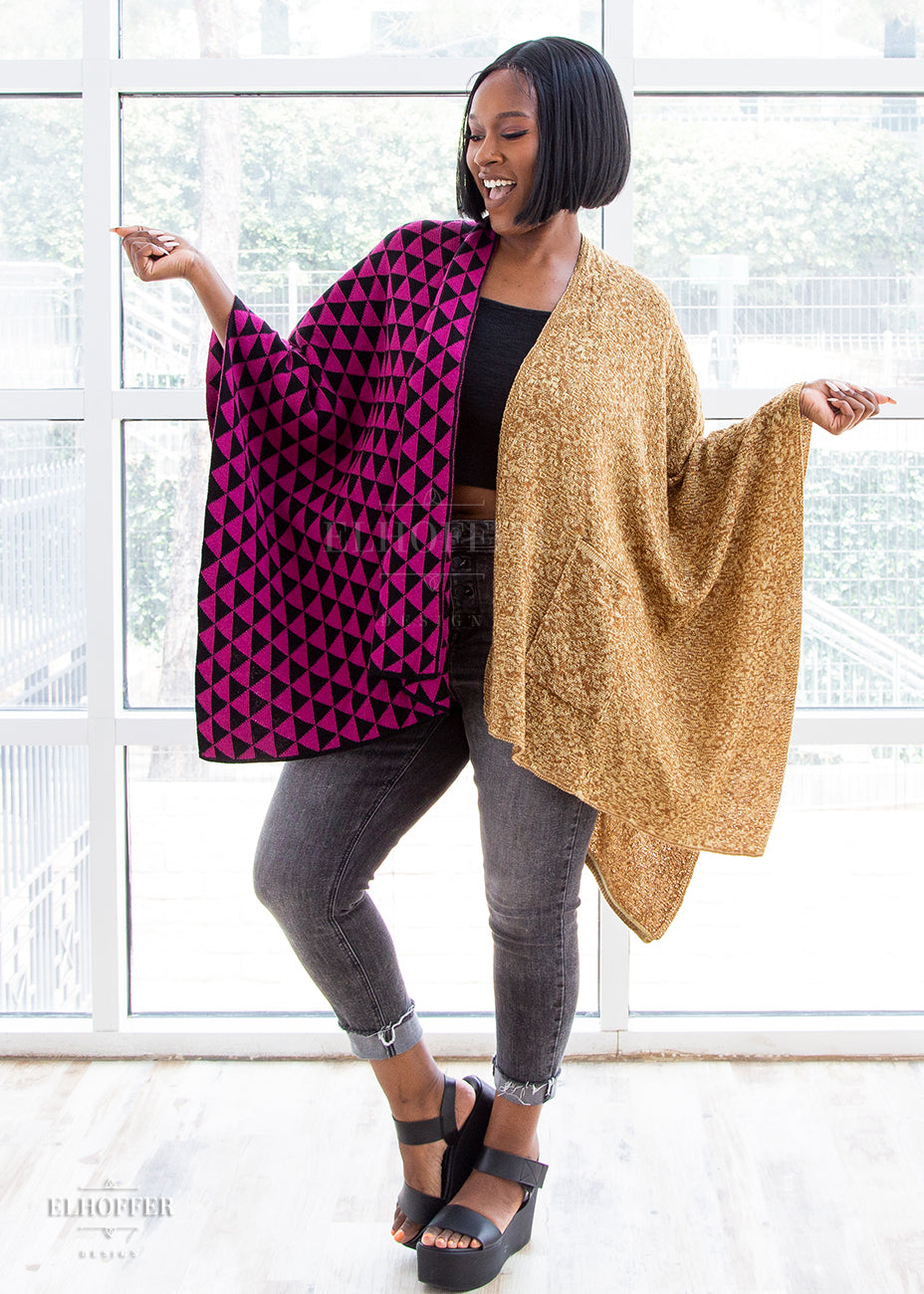 Lynsi stands, holding her arms in a shrug with a smile on her face, showing the poncho across her arms. The right side of the poncho has pink and black triangles and the left side is a golden mixed knit.