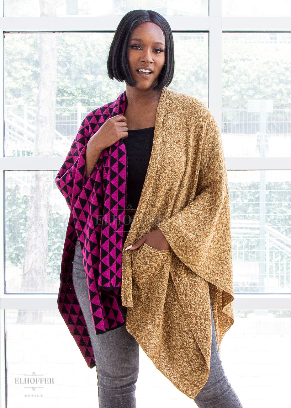 Lynsi, a dark haired dark skinned size L model, wears the poncho with one hand on the collar of the black and pink triangle side and a hand in the pocket of the golden mustard side. She has paired it with a black top and grey jeans.