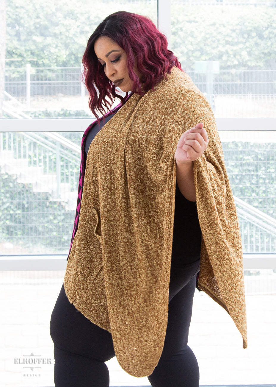 Dawn stands showing the left side of the poncho, a blend of golden and brown tones of yarn and a pocket.