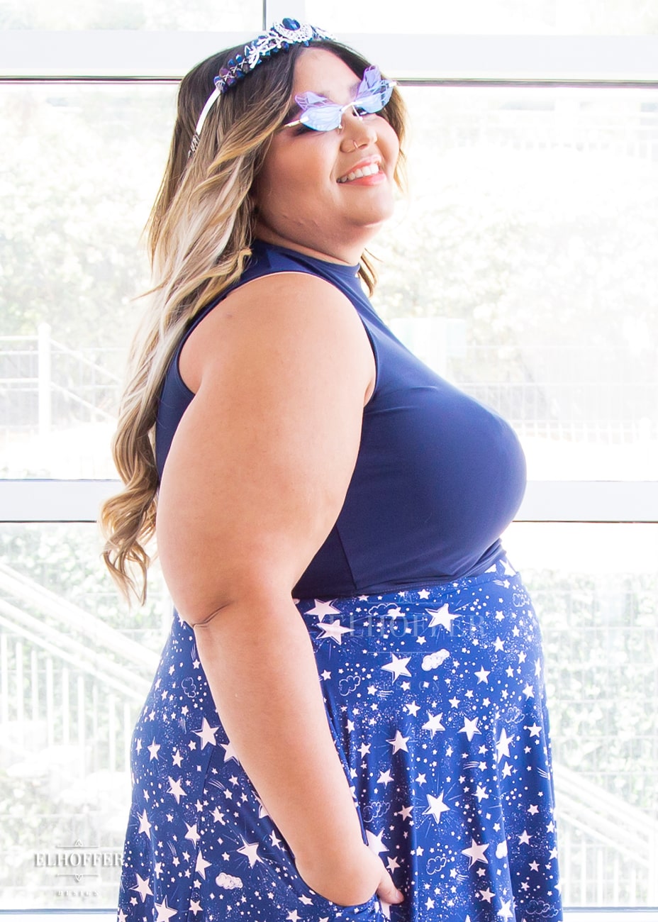 Cori, a medium skinned size 2XL model with brunette and blonde ombre hair, wears a navy a mock turtle necked fitted sleeveless crop top ending at her waist and long skirt covered in a blue and pink celestial print. She stands to the side with her hands in her pocket.