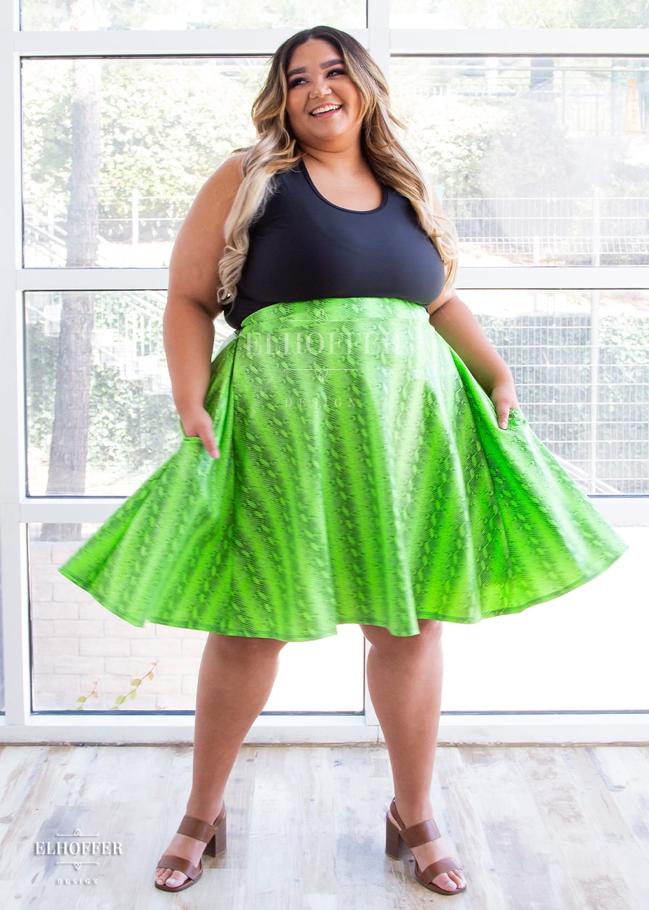 Cori, a medium skinned size 2XL model with brunette and blonde ombre hair, wears a high-waisted knee length full skirt with a fitted matching waistband encased with elastic in the croki green print. The croki green print is a lime green snake print. She is in motion showing off the fullness of the skirt, with her hands in the generous pockets.