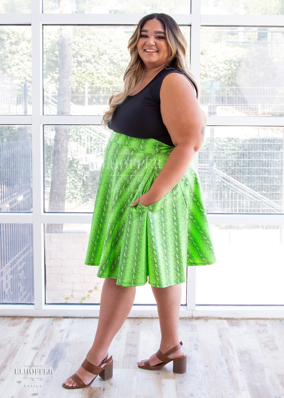 Cori, a medium skinned size 2XL model with brunette and blonde ombre hair, wears a high-waisted knee length full skirt with a fitted matching waistband encased with elastic in the croki green print. The croki green print is a lime green snake print. She stands sideways and shows off the big pockets by placing a hand in one.