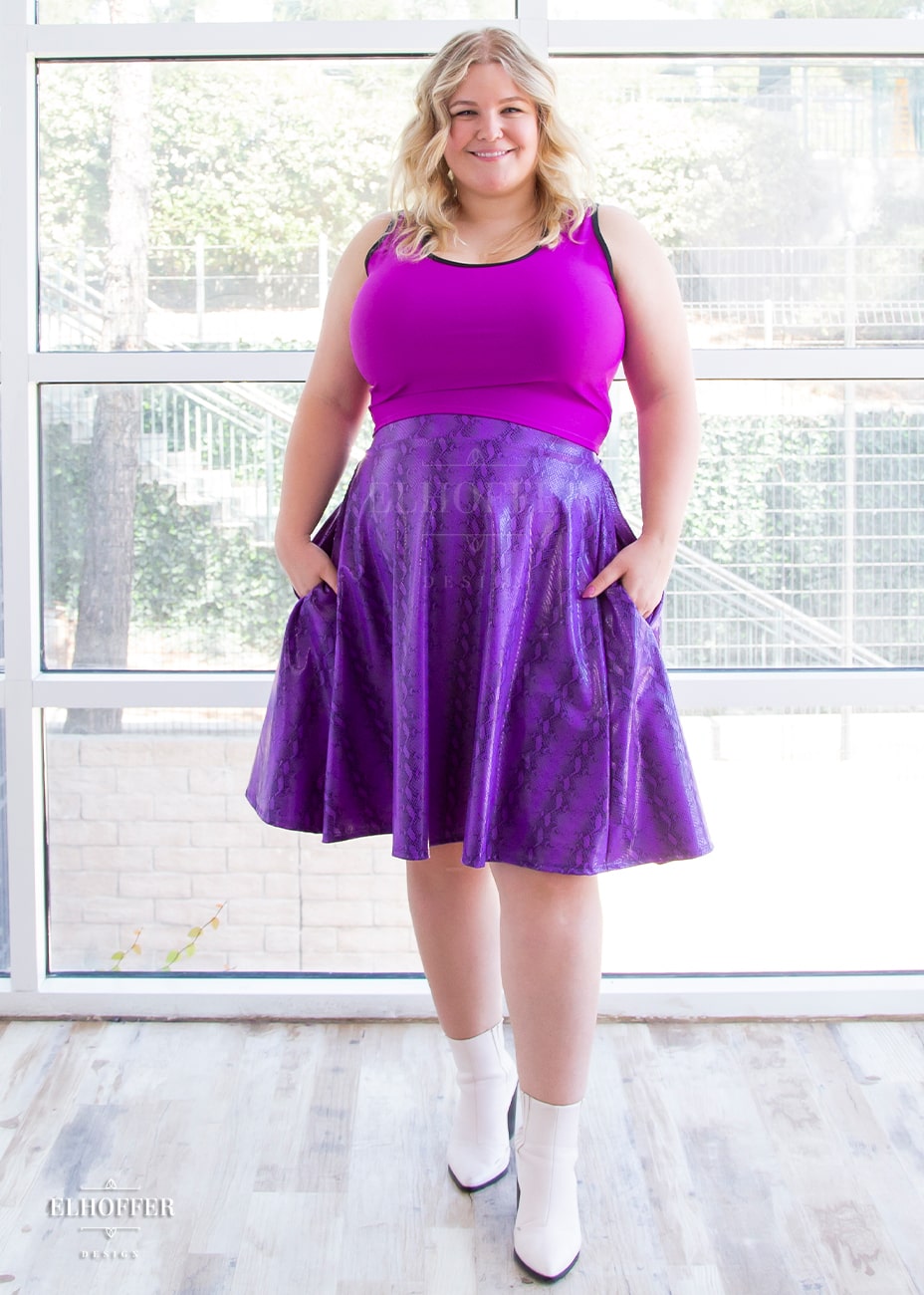 Sarah, a fair skinned size XL model with long blonde hair, wears a high-waisted knee length full skirt with a fitted matching waistband encased with elastic in our purple croki print. The purple croki print is a shiny bright purple snake skin pattern with vertical stripes. She has paired it with a bright magenta Buffy crop tank and white booties.