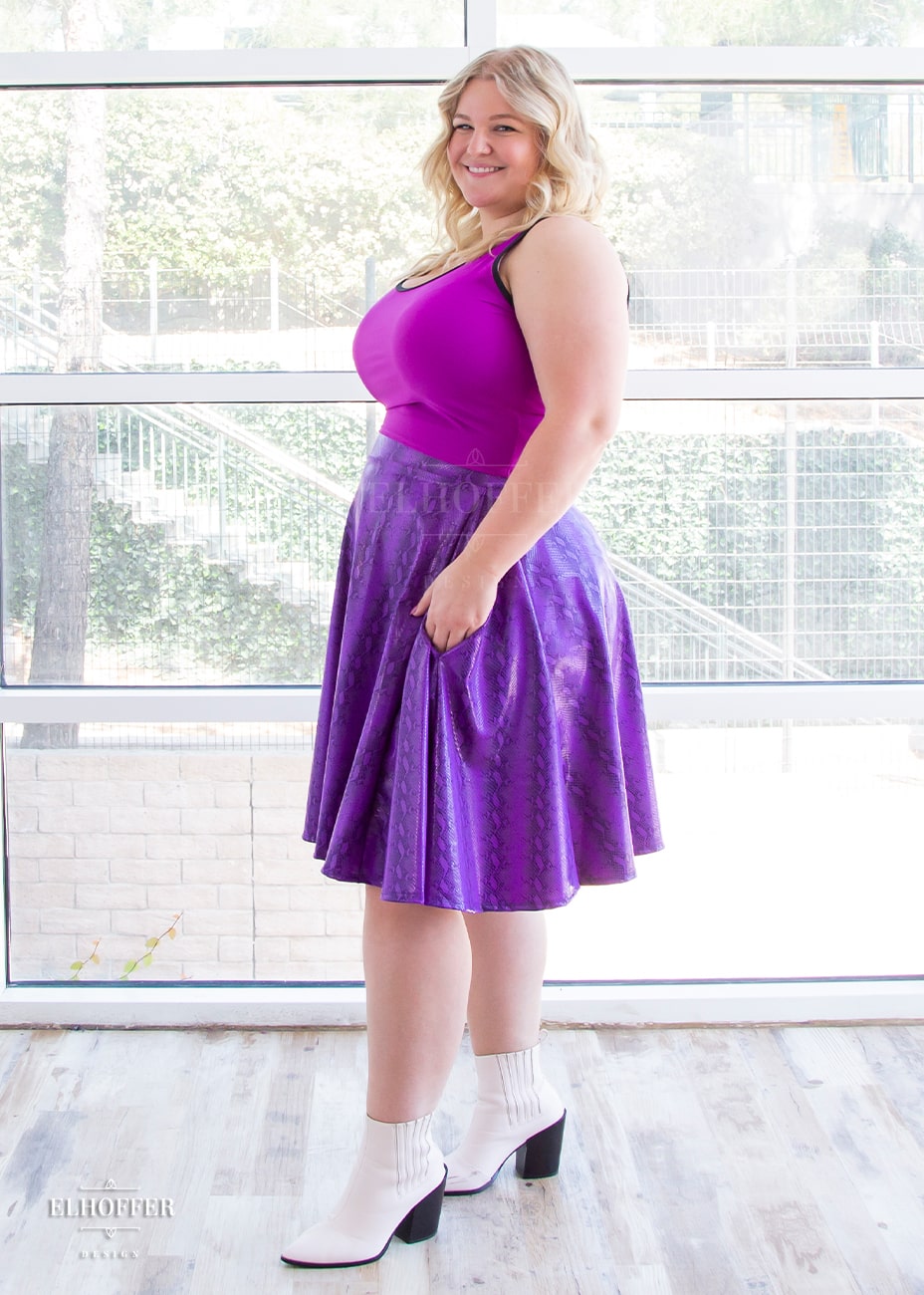 Sarah, a fair skinned size XL model with long blonde hair, wears a high-waisted knee length full skirt with a fitted matching waistband encased with elastic in our purple croki print. The purple croki print is a bright purple shiny snake skin pattern with vertical stripes. She is standing to the side with her hand in one of the generous pockets.