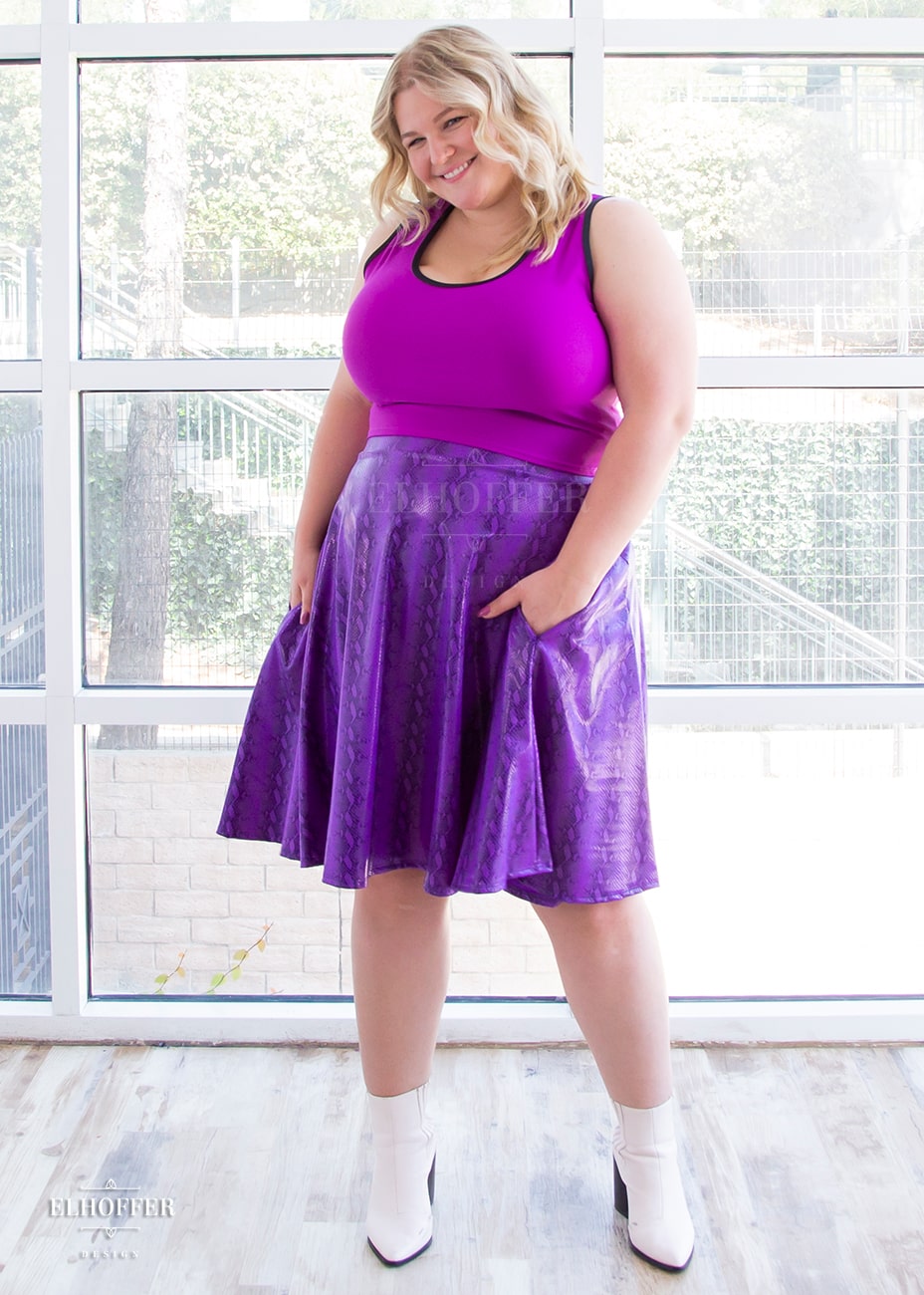 Sarah, a fair skinned size XL model with long blonde hair, wears a high-waisted knee length full skirt with a fitted matching waistband encased with elastic in our purple croki print. The purple croki print is a bright shiny purple snake skin pattern with vertical stripes.