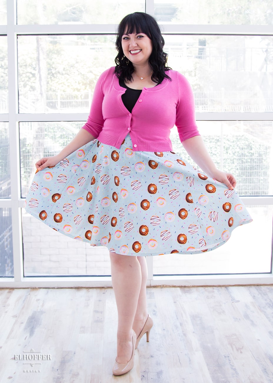 Bernadette, a fair skinned size large model with black hair and bangs, wears a high-waisted knee length full skirt with a fitted matching waistband encased with elastic in our donut stop believing print. The print has a light blue background and is covered in various donuts and sprinkles.