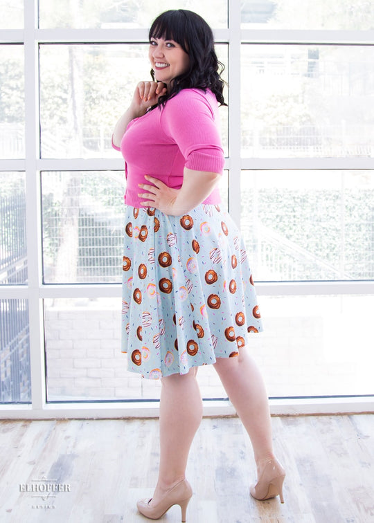 Bernadette, a fair skinned size large model with black hair and bangs, wears a high-waisted knee length full skirt with a fitted matching waistband encased with elastic in our donut stop believing print. The print has a light blue background and is covered in various donuts and sprinkles.