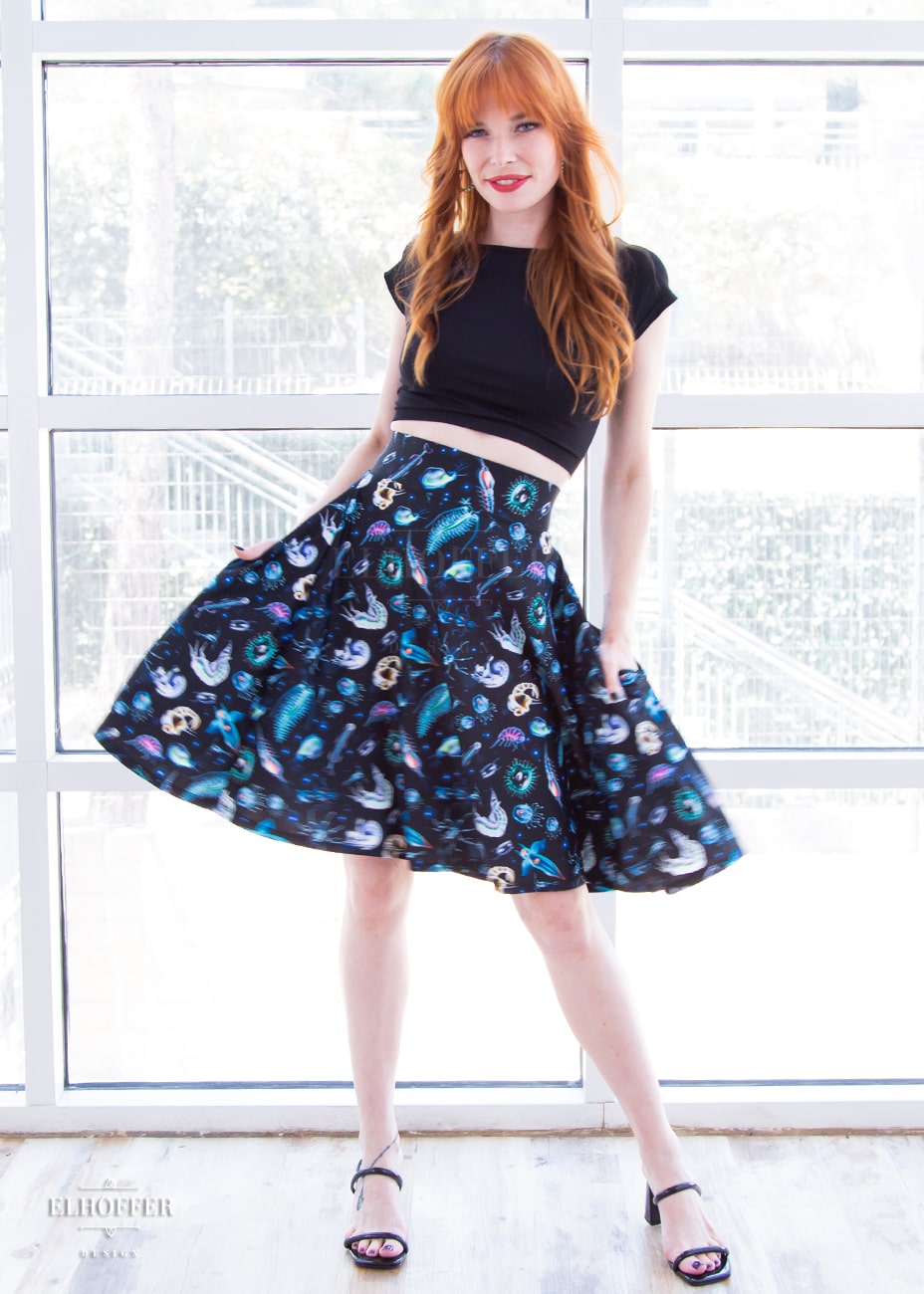 Chloe, a fair skinned size XS model with red hair and bangs, wears a high-waisted knee length full skirt with a fitted matching waistband encased with elastic in our not sea moths print. The print has various small sea creatures on a black background.