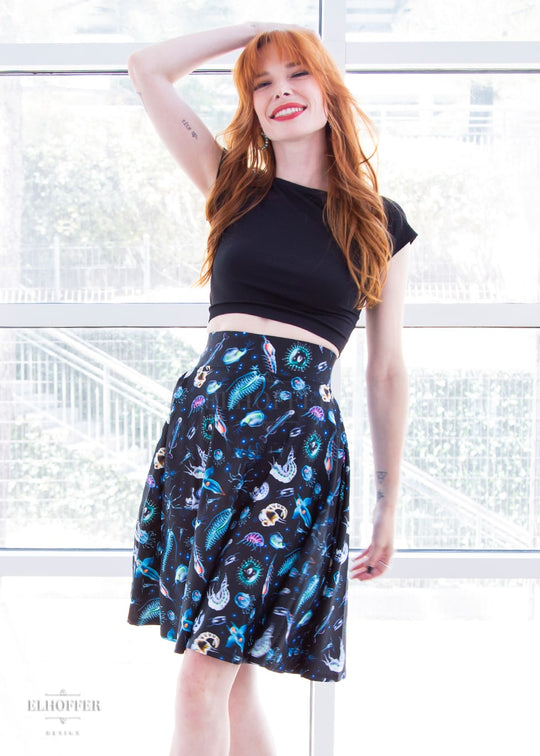 Chloe, a fair skinned size XS model with red hair and bangs, wears a high-waisted knee length full skirt with a fitted matching waistband encased with elastic in our not sea moths print. The print has various small sea creatures on a black background.