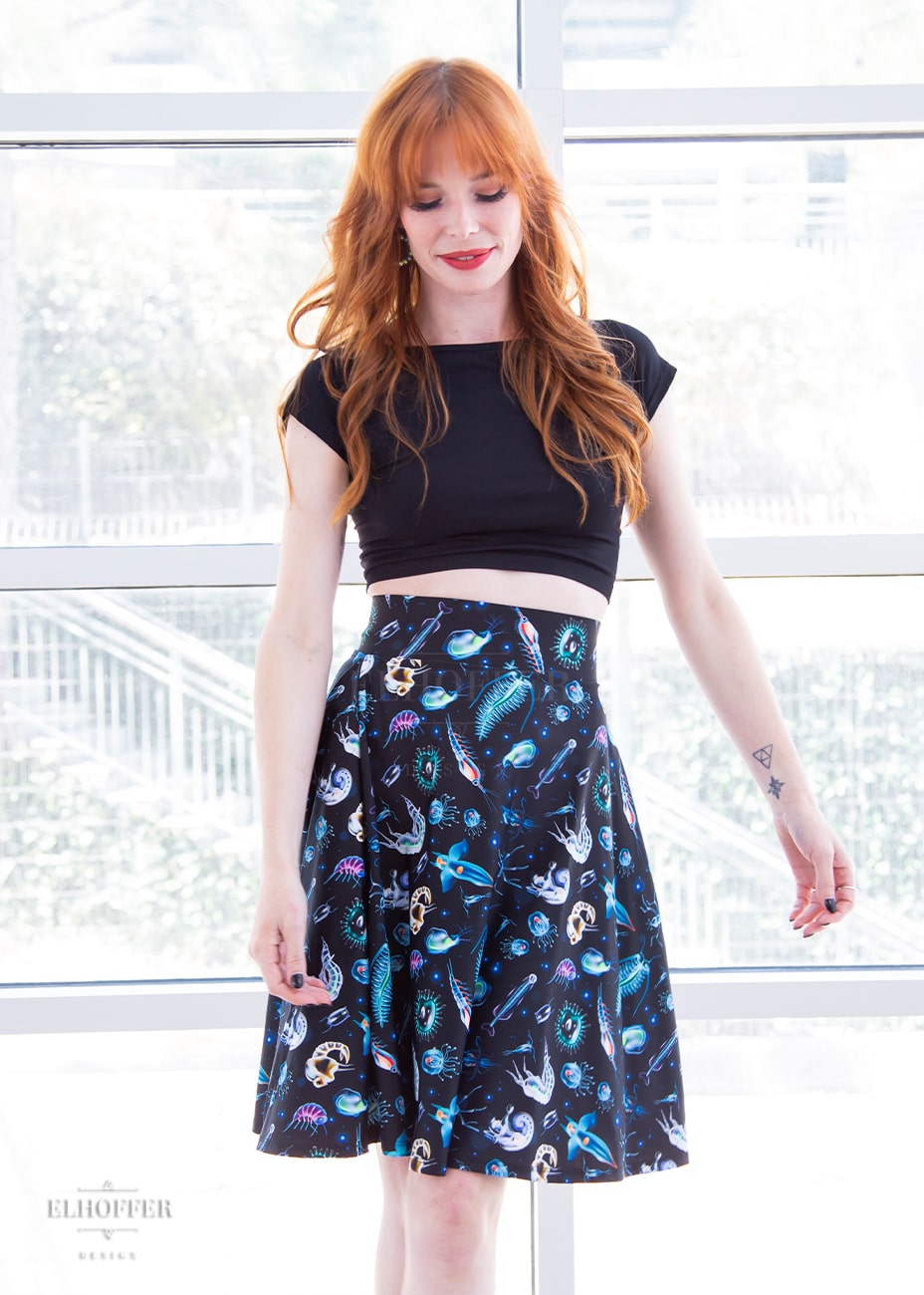 Chloe, a fair skinned size XS model with red hair and bangs, wears a high-waisted knee length full skirt with a fitted matching waistband encased with elastic in our not sea moths print. The print has various small sea creatures on a black background.