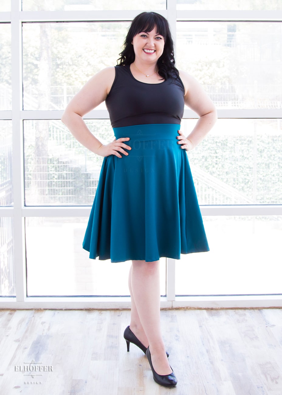 Bernadette, a fair skinned size large model with short black hair and bangs, wears a high-waisted knee length full skirt with a fitted matching waistband encased with elastic in our dark teal ponte.