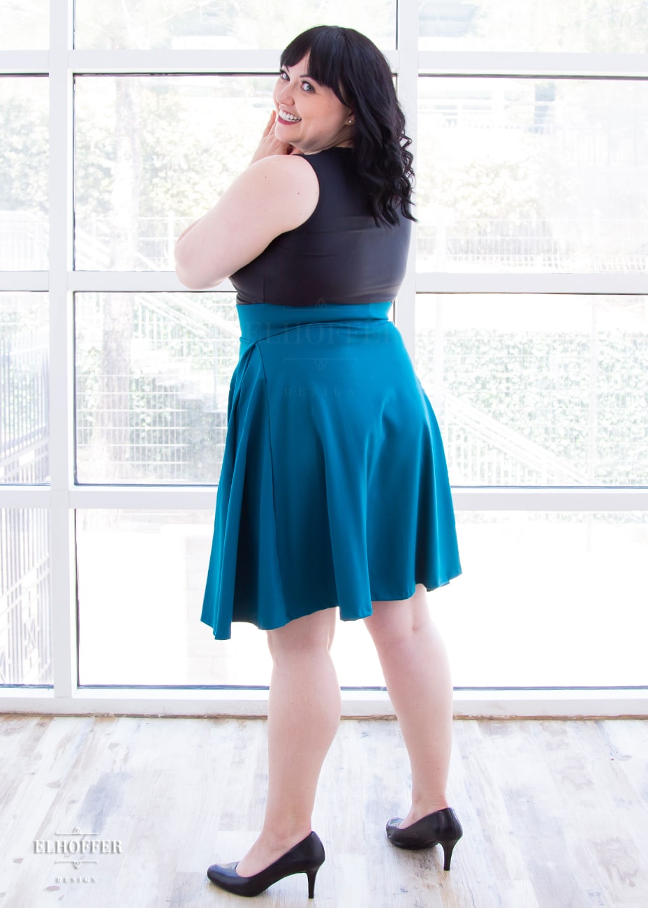Bernadette, a fair skinned size large model with short black hair and bangs, wears a high-waisted knee length full skirt with a fitted matching waistband encased with elastic in our dark teal ponte.
