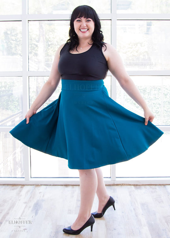 Bernadette, a fair skinned size large model with short black hair and bangs, wears a high-waisted knee length full skirt with a fitted matching waistband encased with elastic in our dark teal ponte.