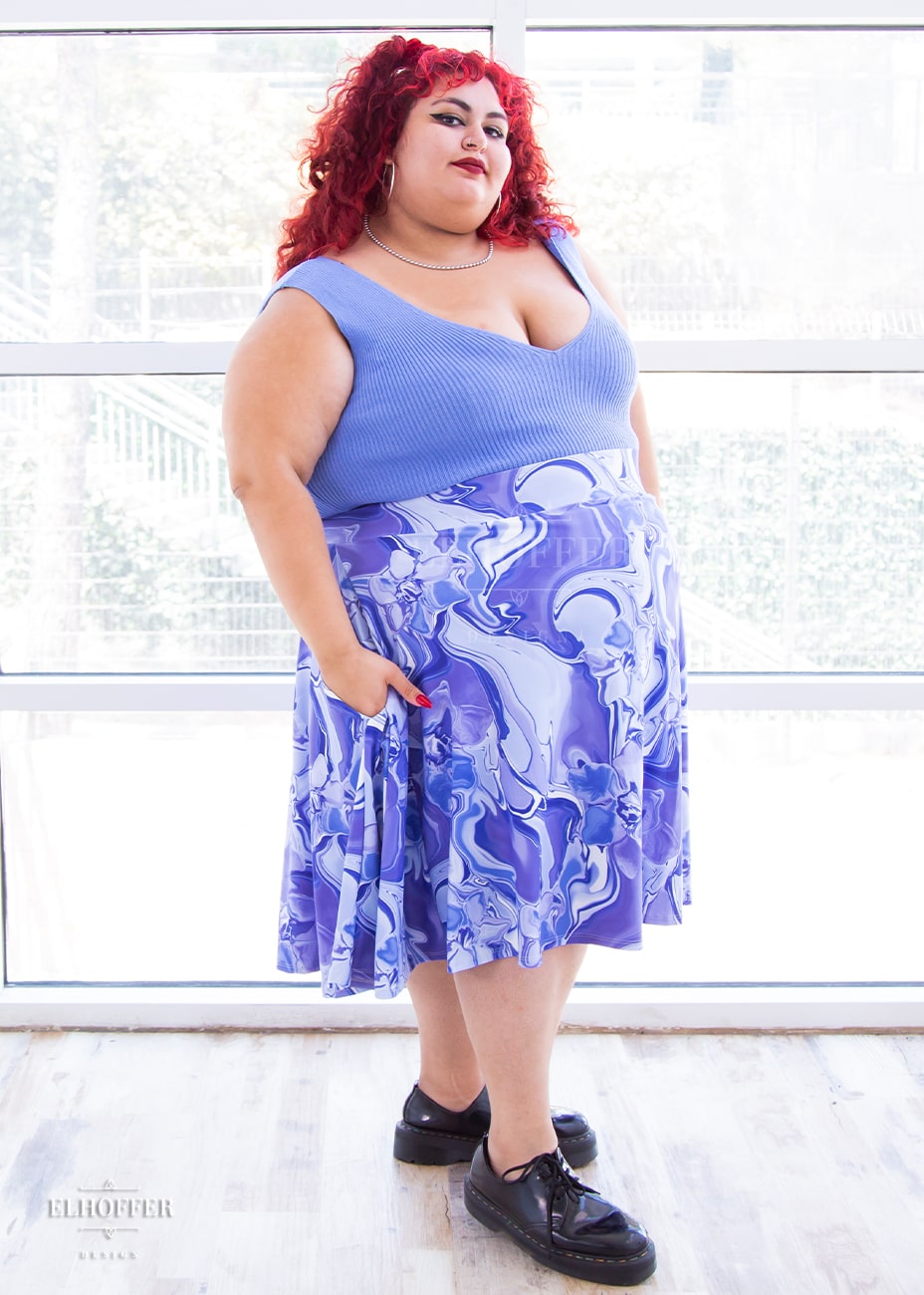 Victoria, an olive skinned 4XL model with curly bright read hair, wears a high-waisted knee length full skirt with a fitted matching waistband encased with elastic in our periwinkle marble print. The periwinkle marble print is a marbled blue, purple, and white pattern all over it.
