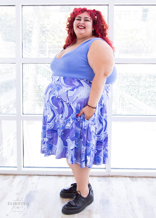 Victoria, an olive skinned 4XL model with curly bright read hair, wears a high-waisted knee length full skirt with a fitted matching waistband encased with elastic in our periwinkle marble print. The periwinkle marble print is a marbled blue, purple, and white pattern all over it.