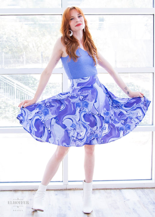 Chloe, a fair skinned size XS model with red hair and bangs, wears a high-waisted knee length full skirt with a fitted matching waistband encased with elastic in our periwinkle marble print. The periwinkle marble print is a marbled blue, purple, and white pattern all over it.