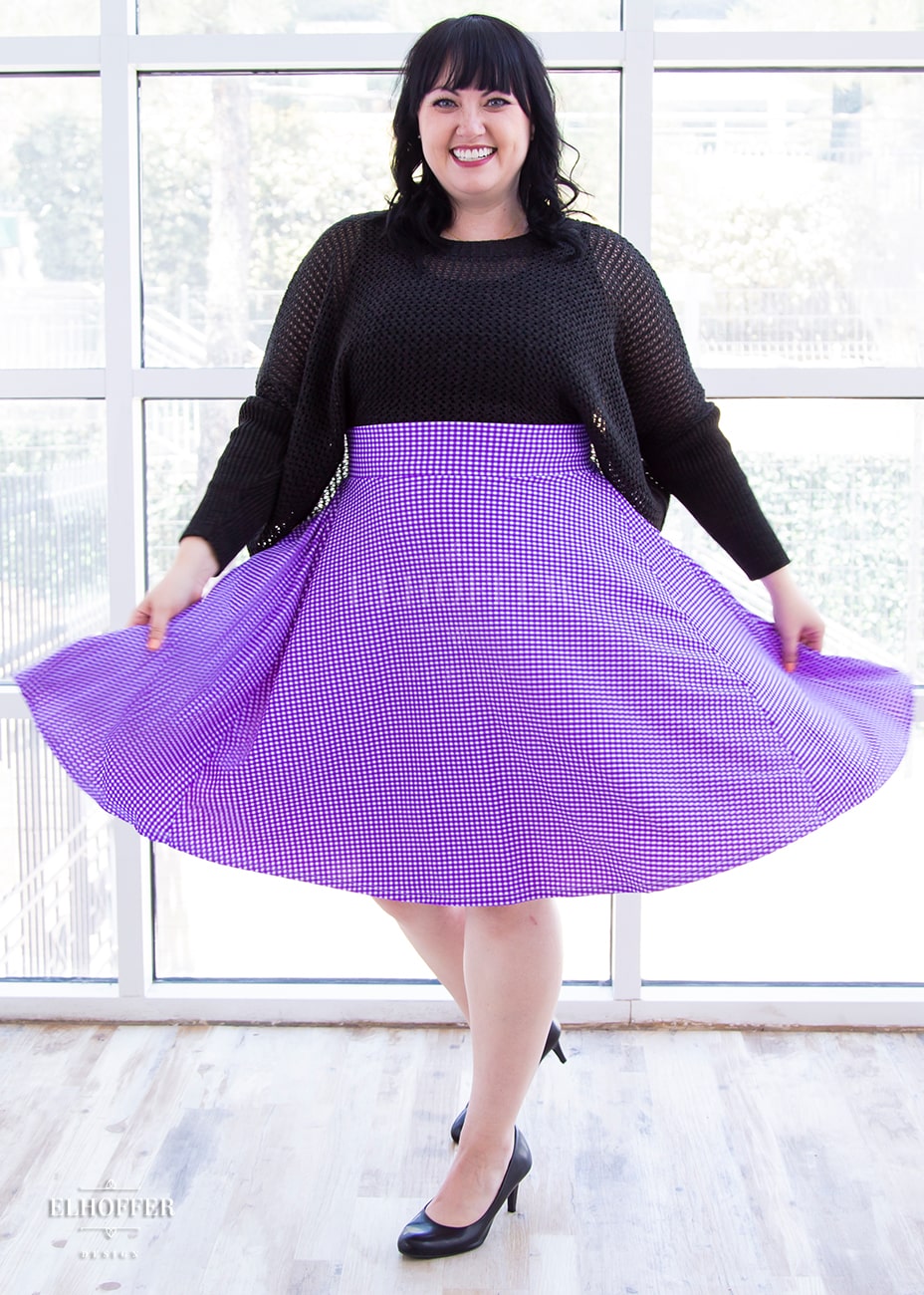 Bernadette, a size large model with short black hair and bangs, wears a high-waisted knee length full skirt with a fitted matching waistband encased with elastic in our grape gingham. The grape gingham print is a bright purple gingham.