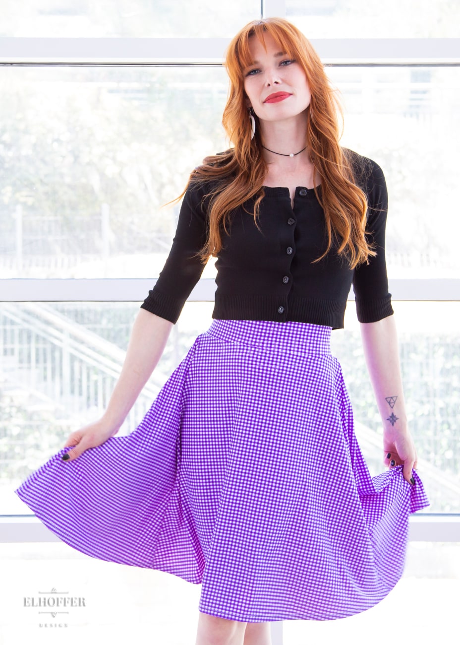 Chloe, a fair skinned size XS model with red hair and bangs, wears a high-waisted knee length full skirt with a fitted matching waistband encased with elastic in our grape gingham. The grape gingham print is a bright purple gingham.