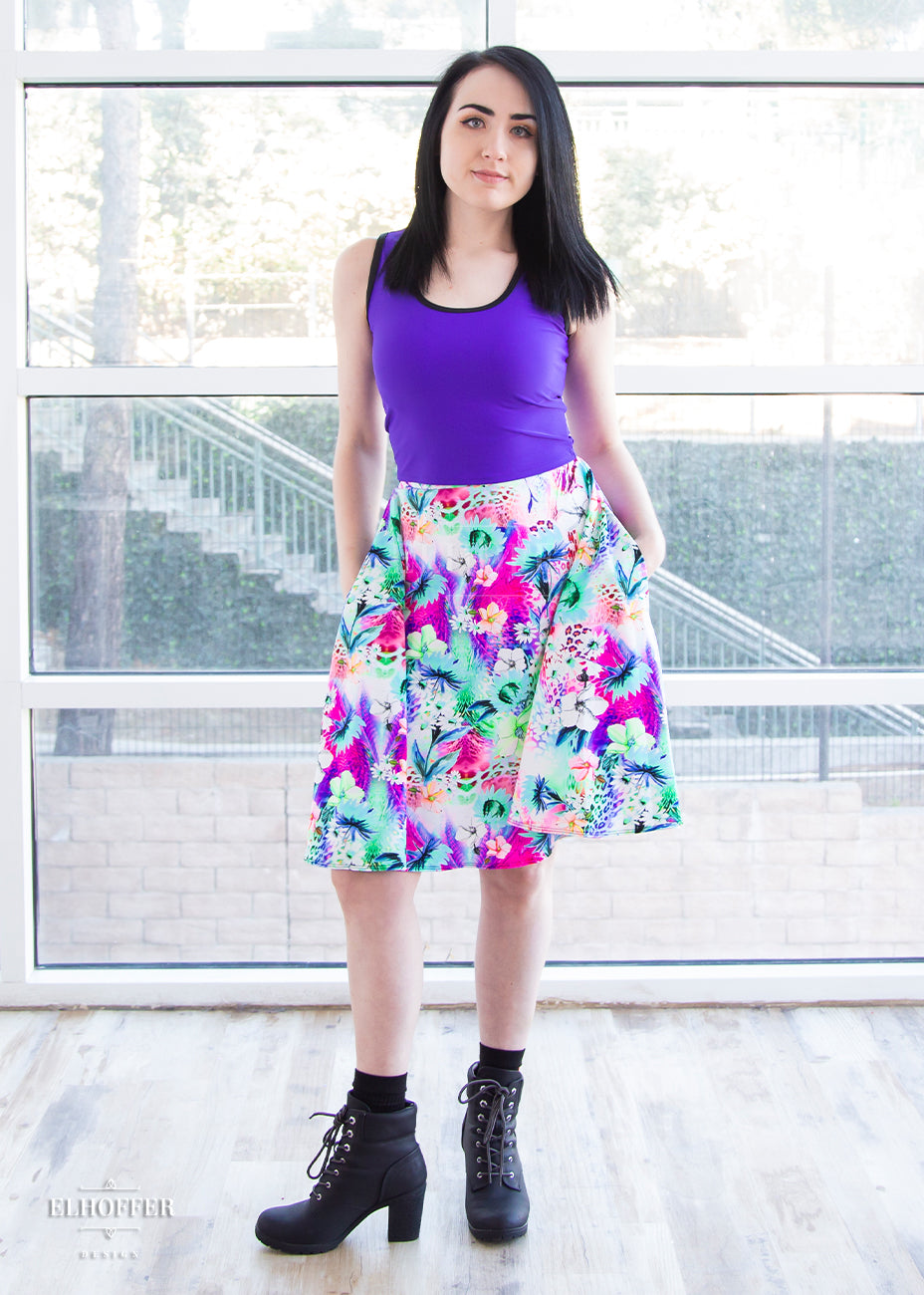 Amy, a fair skinned size small model with short dark hair, wears a high-waisted knee length full skirt with a fitted matching waistband encased with elastic in our rainbow jungle. The pattern is bright blue, pink and green with tropical flowers.