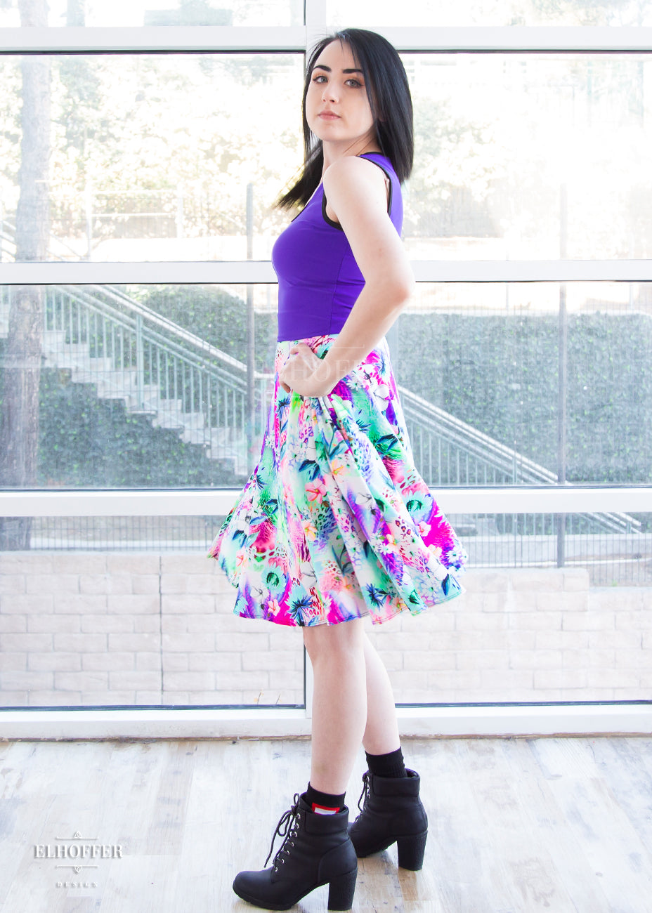 Amy, a fair skinned size small model with short dark hair, wears a high-waisted knee length full skirt with a fitted matching waistband encased with elastic in our rainbow jungle. The pattern is bright blue, pink and green with tropical flowers.