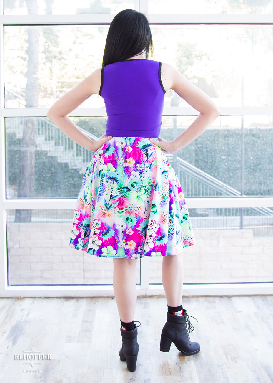 Amy, a fair skinned size small model with short dark hair, wears a high-waisted knee length full skirt with a fitted matching waistband encased with elastic in our rainbow jungle. The pattern is bright blue, pink and green with tropical flowers.