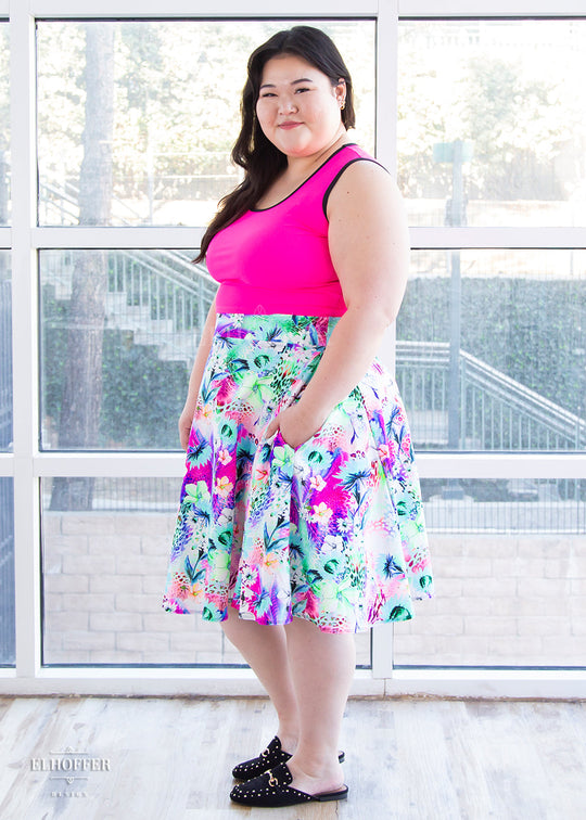 Ashley, a fair skinned size XL model with long brown hair, wears a high-waisted knee length full skirt with a fitted matching waistband encased with elastic in our rainbow jungle. The pattern is bright blue, pink and green with tropical flowers.