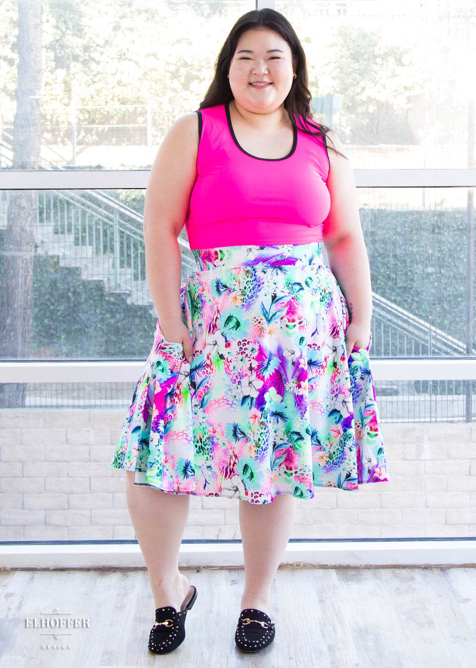 Ashley, a fair skinned size XL model with long brown hair, wears a high-waisted knee length full skirt with a fitted matching waistband encased with elastic in our rainbow jungle. The pattern is bright blue, pink and green with tropical flowers.